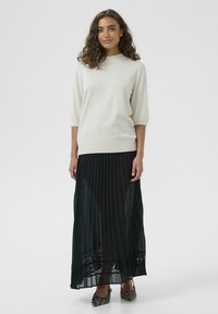 Svetlá krémová pletená blúzka s krátkymi rukávmi, spojená s dlhou čiernou plisovanou sukňou s čipkovým lemom na okraji, nosená s pruhovanými lodičkami na vysokom opätku.