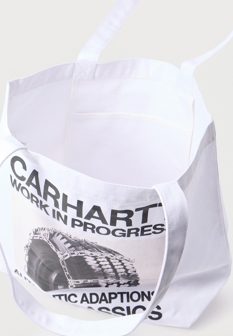 Sac fourre-tout blanc avec un texte noir « Carhartt Work In Progress » et un pneu enroulé dans des chaînes imprimé sur un côté.