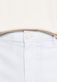 Detailní záběr na světle modré kalhoty s knoflíkovým zapínáním a s béžovou košilí zasunutou v pase.