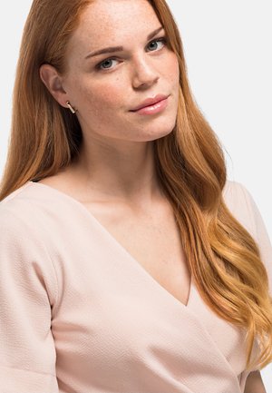 Top avvolgente rosa chiaro in tessuto strutturato, con scollatura a V profonda. Lunghi capelli rossi ondulati che cadono su una spalla. Semplici orecchini d'oro.