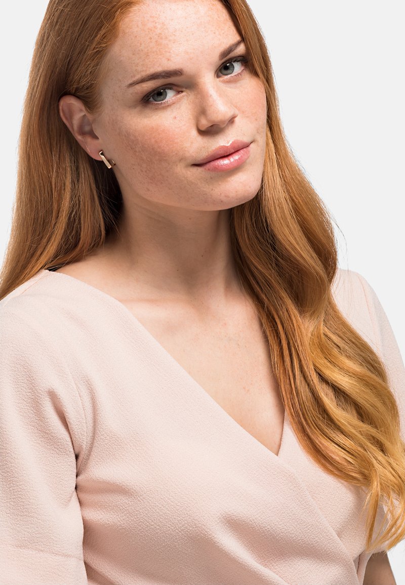 Haut enveloppant rose clair en tissu texturé, avec un décolleté en V profond. Cheveux longs et ondulés roux tombant sur l'épaule. Boucles d'oreilles en or simples.