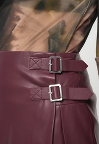 Μαρούλι δερμάτινη φούστα με πτυχώσεις και δύο ασημένιες μεταλλικές αγκράφες. Η φούστα διαθέτει αντιθετική ραφή και ομαλή υφή.