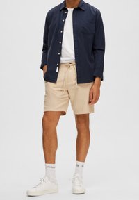 Homme portant une chemise bleu marine à boutons par-dessus un t-shirt blanc, un short beige avec cordon, des chaussettes blanches et des baskets blanches, debout devant un fond uni.