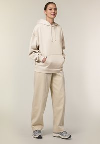 Felpa oversize beige con tasca frontale e cordino, abbinata a pantaloni beige larghi e scarpe da ginnastica grigie. Tessuto dalla texture morbida.