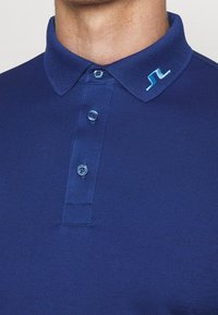 Námořnická modrá polokošile s žebrovaným límcem, třemi knoflíky a malým logem světle modré barvy na límci. Hladká textura, ležérní design.