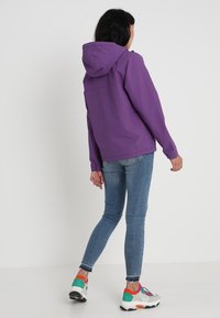 Lilla, let jakke med hætte, der har en glat tekstur og elastiske manchetter, båret over lyseblå skinny jeans og farverige sneakers.