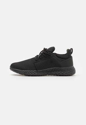 Black athletic shoe with a mesh upper, round toe, laces, and a textured black rubber sole. Features a pull tab on the heel.