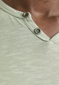Lys grøn henley skjorte med V-hals, to metalliske knapper på kraven og et blødt, tekstureret stof med et subtilt mønster.