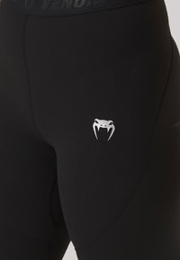 Fekete leggings, rugalmas anyagból, ezüst színű fang logóval a comb felső részén és texturált, márkázott derékpánttal.