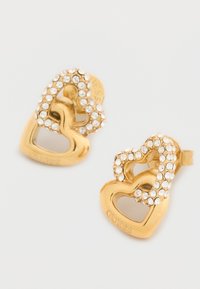 Σκουλαρίκια σε σχήμα καρδιάς με χρυσό τόνο, διακοσμημένα με καθαρά στρας που περιγράφουν ένα κενό κέντρο. Το όνομα της μάρκας "GUESS" είναι χαραγμένο στο πλάι.