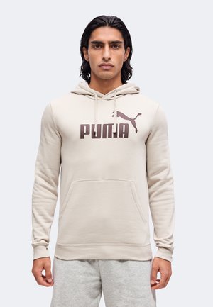 Homme aux cheveux foncés portant un sweat à capuche beige Puma et un pantalon de survêtement gris clair, debout devant un fond clair uni.