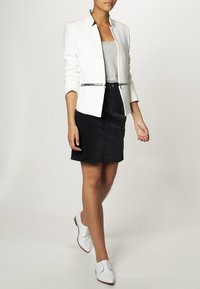 Weißer Blazer mit einem scharfen Kragen, tailliertem Design und schwarzem Reißverschluss, kombiniert mit einem grauen Tanktop und einem schwarzen Rock, abgerundet mit weißen Schuhen.
