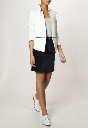Blazer blanc avec un col pointu, design ajusté et détails de fermeture éclair noire, associé à un débardeur gris et une jupe noire, complété par des chaussures blanches.
