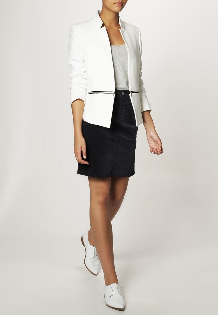 Weißer Blazer mit einem scharfen Kragen, tailliertem Design und schwarzem Reißverschluss, kombiniert mit einem grauen Tanktop und einem schwarzen Rock, abgerundet mit weißen Schuhen.
