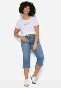 Balta marškinėliai su „Capri“ užrašu, šviesiai mėlyni capri tipo džinsai, žalias diržas ir pilki sportbačiai. Modelis stovi pozavime su viena ranka ant klubo.