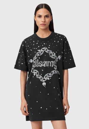 Giovane donna che indossa un vestito a t-shirt nero largo con stampe di catene e gemme argentate, in piedi davanti a uno sfondo grigio chiaro uniforme.