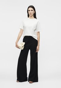 Femme aux longs cheveux foncés portant un haut à manches courtes blanc, un pantalon noir à jambes larges, des sandales noires et tenant une petite pochette beige.