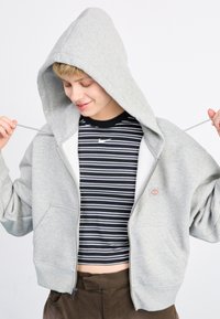 Felpa grigia con zip e cappuccio ampio, due tasche anteriori e polsi a coste, abbinata a una maglietta corta a righe bianche e nere sotto.