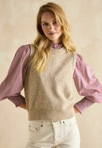 Beige gestrickte Weste über einer bordeaux- und weiß gestreiften Bluse mit Rüschensaum am Kragen und den Ärmelmanschetten; kombiniert mit weißen Jeans.