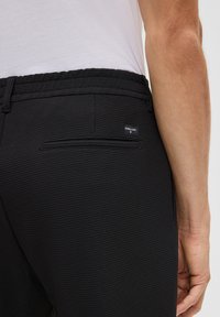 Zbliżenie na osobę noszącą teksturowane czarne spodnie z tylną kieszenią i małą metką marki, w połączeniu z białą koszulą włożoną w spodnie.