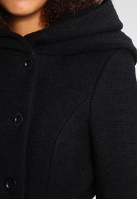 Cappotto in misto lana nera con un ampio cappuccio, tessuto testurizzato e chiusure a bottone. Il colletto è largo, conferendo un aspetto strutturato.