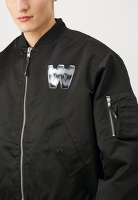 Čierna bomber bunda so zipsom vpredu, ryhovaným golierom a veľkým strieborným logom na hrudi. Obsahuje bočné a zipsové vrecká.