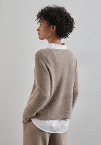 Beige, fluffig stickad tröja med ribbad textur, med rund hals och sänkt axel, bärs över en vit skjorta med krage.