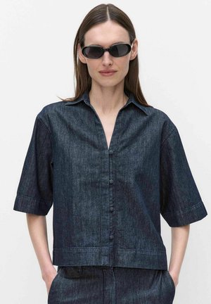 Nainen, jolla on yllään tummansininen lyhythihainen nappipaita ja samansävyiset housut sekä mustat ovaalit aurinkolasit, seisoo yksivärisen valkoisen taustan edessä.