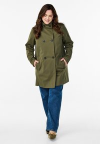 Cappotto doppiopetto verde oliva con bottoni neri, tasche laterali e colletto alto, abbinato a jeans in denim blu e scarpe verdi.