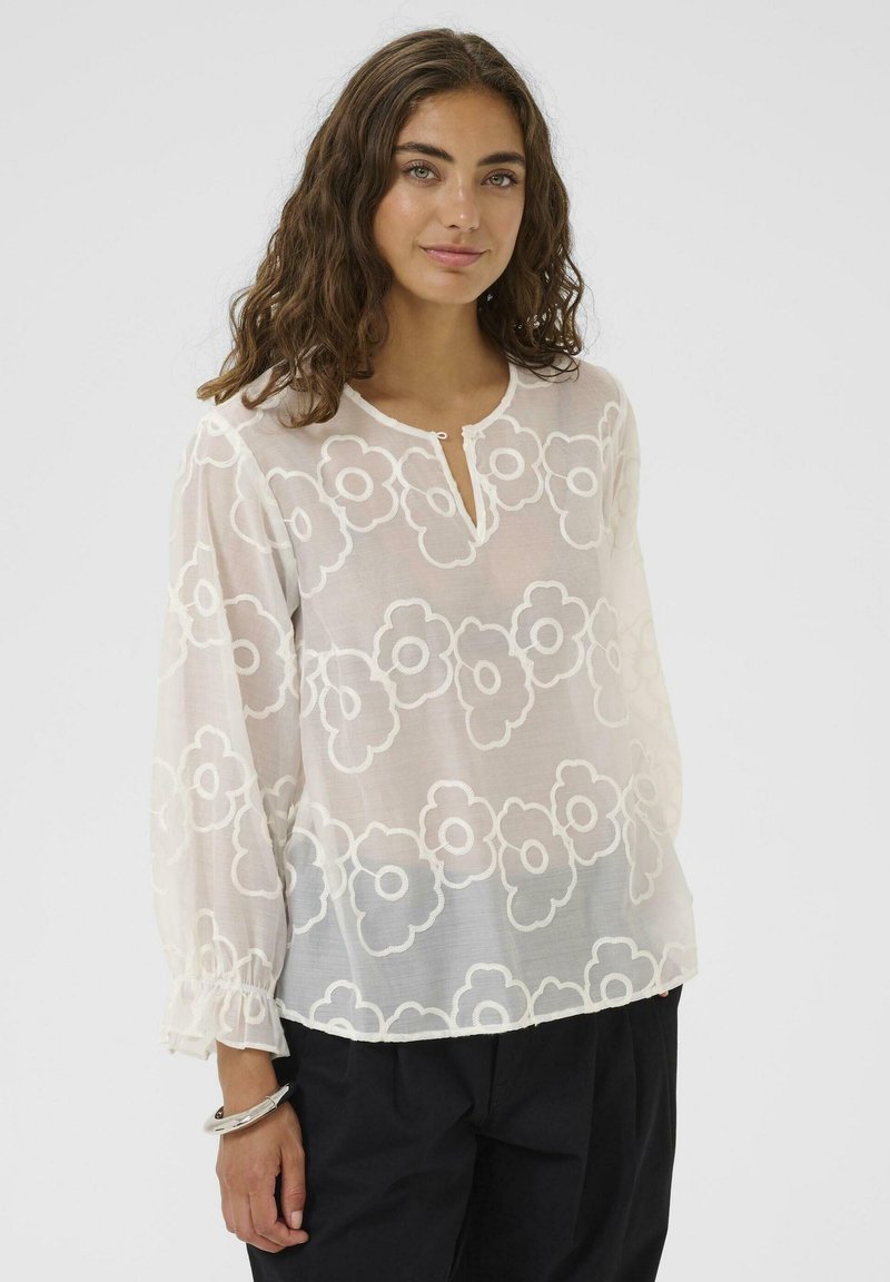 Chemisier blanc transparent avec broderies florales, présentant un col boutonné et des poignets froncés pour plus de détail et de texture.