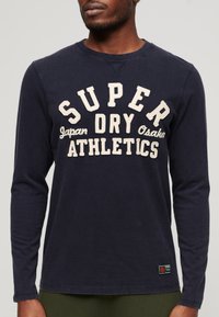 Mørkeblå langærmet t-shirt lavet af blød bomuld. Har et hvidt broderet logo "SUPER DRY ATHLETICS" på forsiden og et brandet mærke ved kanten.