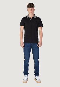 Camisa polo negra con un cuello gris, mangas cortas y un logo en el pecho, combinada con jeans azules y zapatillas blancas con acentos verdes.