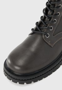 Bottine en cuir gris foncé avec un bout arrondi, semelle en caoutchouc noir robuste et lacets bien visibles. Présente des accents de couture visibles.