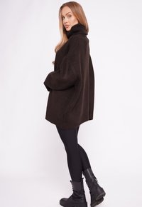 Übergroßer dunkelbrauner Strickpullover mit hohem Kragen, getragen über schwarzen Leggings und schwarzen Stiefeln. Ausgeprägte Textur und entspannte Passform.