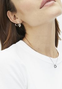 Des boucles d'oreilles en argent en différentes tailles et un délicat collier en chaîne avec un petit pendentif en forme de cœur, porté sur un t-shirt blanc.
