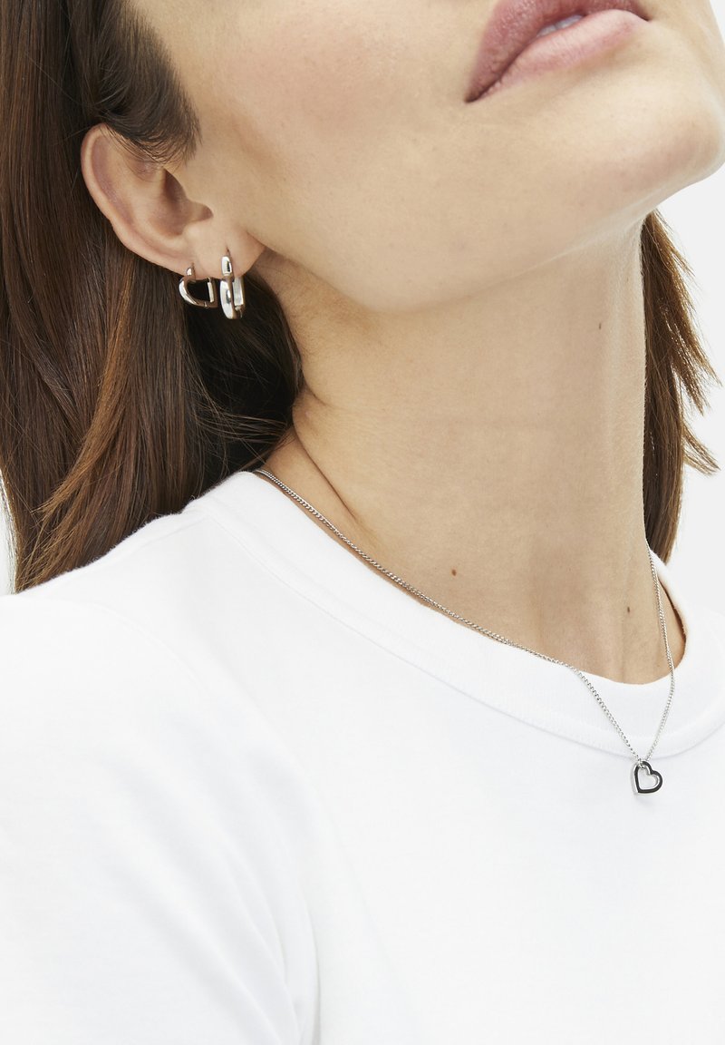Des boucles d'oreilles en argent en différentes tailles et un délicat collier en chaîne avec un petit pendentif en forme de cœur, porté sur un t-shirt blanc.