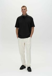 Sort bomulds t-shirt, løs pasform, rund hals; cremefarvede bukser, lige ben; sorte sko; mønstret tørklæde om halsen. Enkel baggrund.