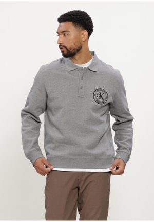 Man draagt een grijs poloshirt met lange mouwen en een rond Calvin Klein-logo op de borst, houdt de zoom vast en kijkt naar links.
