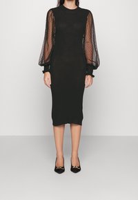 Robe noire en midi avec un corsage ajusté et des manches bouffantes transparentes à pois. Associée à des escarpins noirs avec un accent doré décoratif.