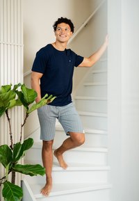 Námořnické bavlněné tričko a šedé vzorované šortky, které nosí osoba na schodech, s blízkou zelenou rostlinou v květináči a bílými zdmi.