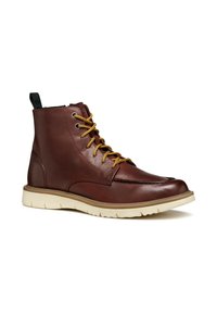 Botas de tobillo de cuero marrón con cordones amarillos, ojales grises y una lengüeta negra. Cuentan con una suela de goma blanca con un acabado texturizado.