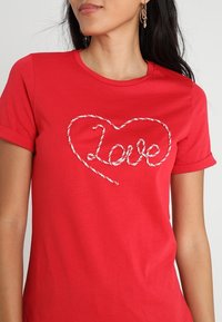 Camiseta de algodón roja con un diseño de script texturizado en blanco que dice "amor" dentro de una forma de corazón. Mangas cortas y cuello redondo.