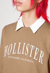 Brun sweatshirt med vit krage, med texten "HOLLISTER" och "SOUTHERN CALIFORNIA" nedanför, mjuk textur, avslappnad design.