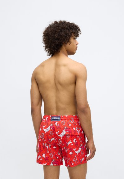 Jaunas vyras su garbanotais plaukais stovi atsukęs nugarą, dėvi raudonus maudymosi šortus su baltais jūrų gyvūnų raštais, priešais paprastą baltą foną.