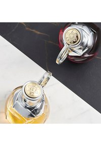 Twee parfumflessen worden getoond, één met een heldere glazen bodem en een zilveren dop met gouden accenten, de andere met een donkerrode bodem en een vergelijkbare dop.