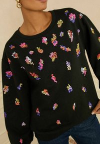 Melns džemperis ar krāsainiem sekunžu rotājumiem dažādās formās un izmēros. Iezīmē klasisku apkakli un vaļīgas piedurknes.