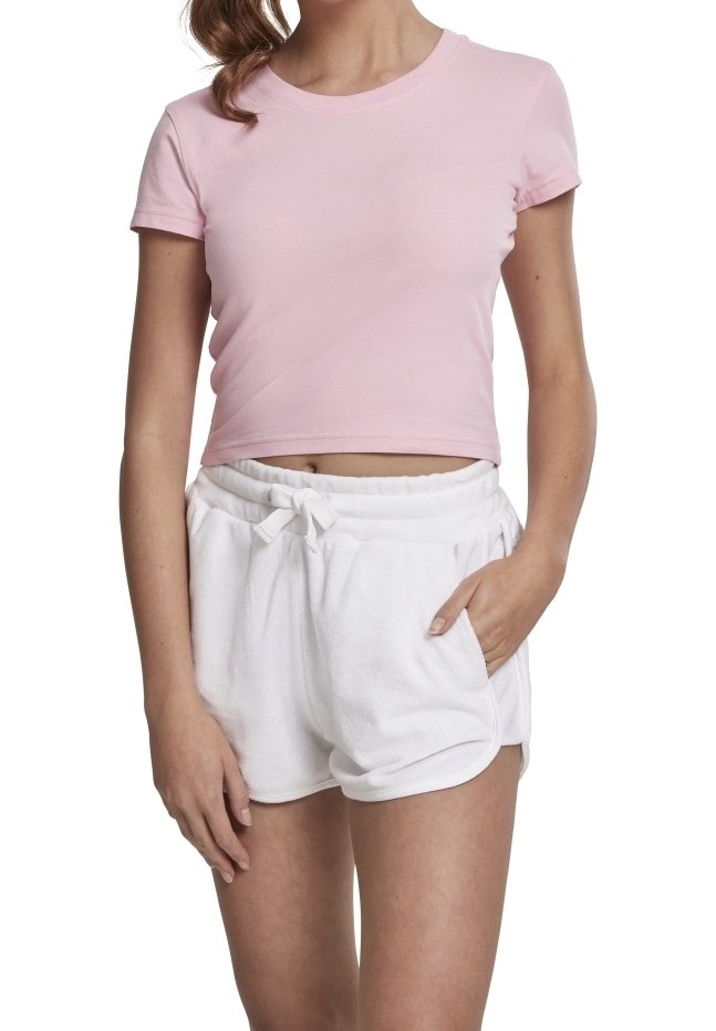 Femme portant un t-shirt rose court et un short blanc à cordon avec une main dans la poche, debout sur un fond blanc.