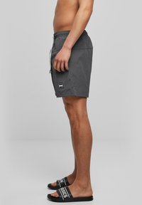 Grå badeshorts med elastisk midje og snor; side logo-merking. Kombinert med svarte sandaler med hvit tekst på remmene.