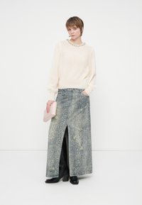 Nainen, jolla on päällään kermanvärinen neule, jossa on koristeltu kaulus, pitkä käärmeennahkakuosiset hame, jossa on edessä halkio, mustat saappaat ja hän pitää vaaleanpunaista clutch-laukkua.