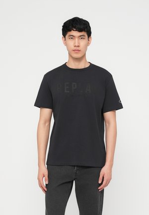 Ung mann med kort svart hår, iført en svart Replay T-skjorte og mørke jeans, står mot en ensfarget hvit bakgrunn.